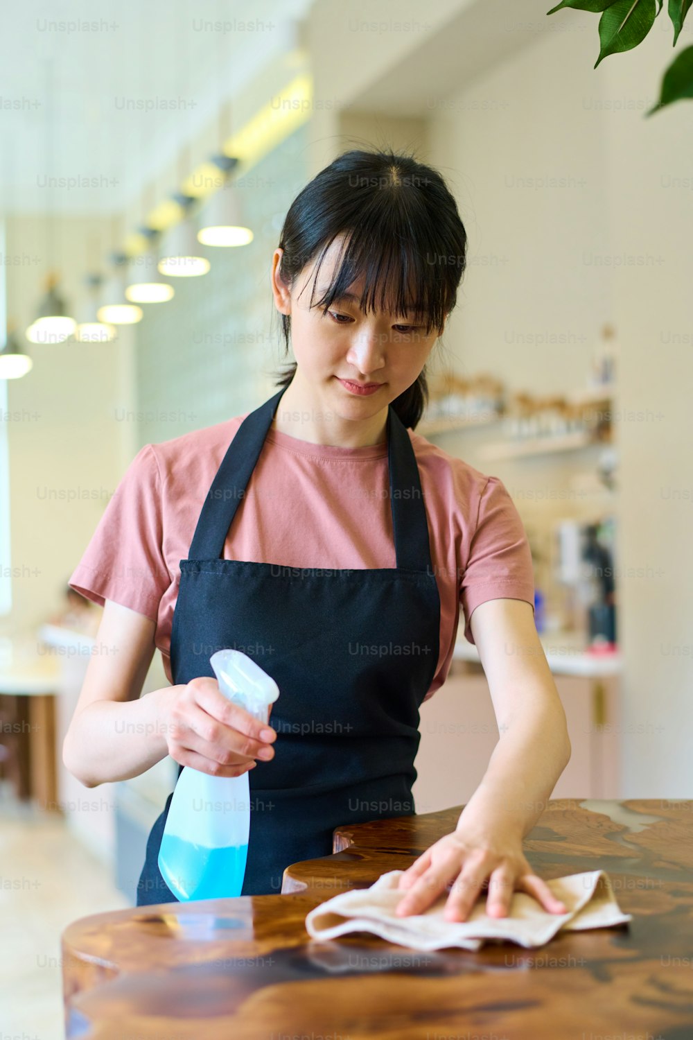 Joven camarera asiática en delantal limpiando la mesa con un agente de limpieza antes de la apertura de la cafetería