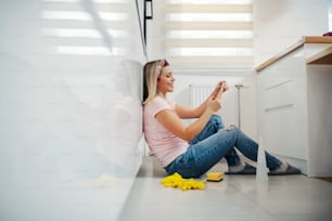 A woman sits in kitchen, taking a break from chores and texting messages on the phone.