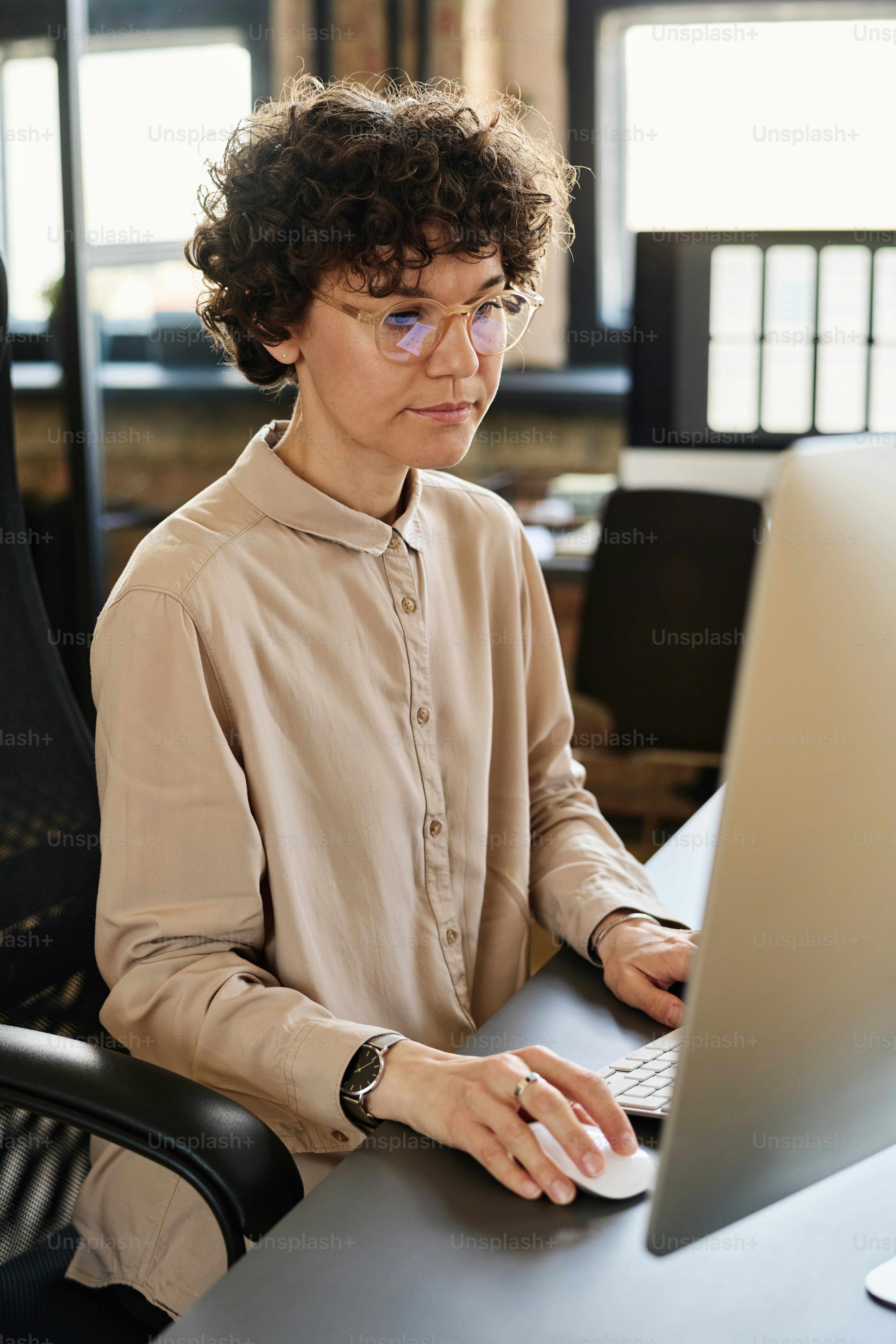 Office employee in eyeglasses sitting at her workplace at office and working on computer using mouse and key