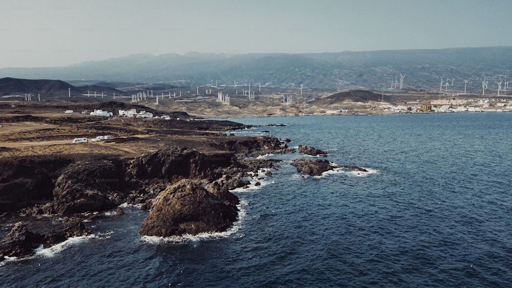 Luftaufnahme der Küste von Felsen und Meereswellen mit kleiner Stadt im Hintergrund. Blauer Himmel und Meereswellen mit Windmühlen und Bergen. Konzept des Reisens und des landschaftlichen Ortes