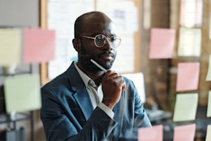 Homme d’affaires africain sérieux en lunettes debout devant un mur de verre avec des notes et la lecture de plans d’affaires