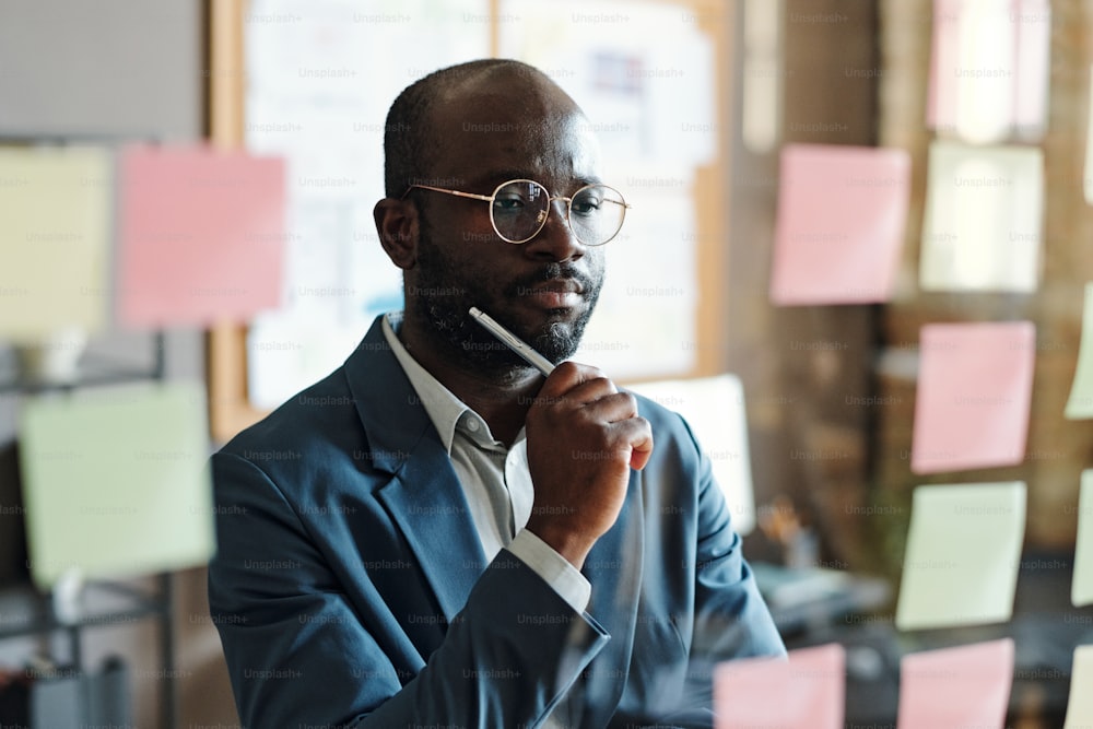 Serious African businessman in eyeglasses standing in front of glass wall with notes and reading business plans