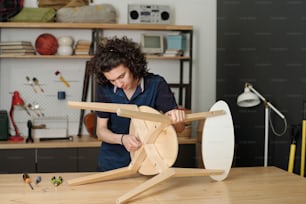 Adolescent contemporain en tenue décontractée se penchant sur une table tout en réparant ou en fabriquant une chaise en bois et en utilisant un outil à main pour fixer les détails