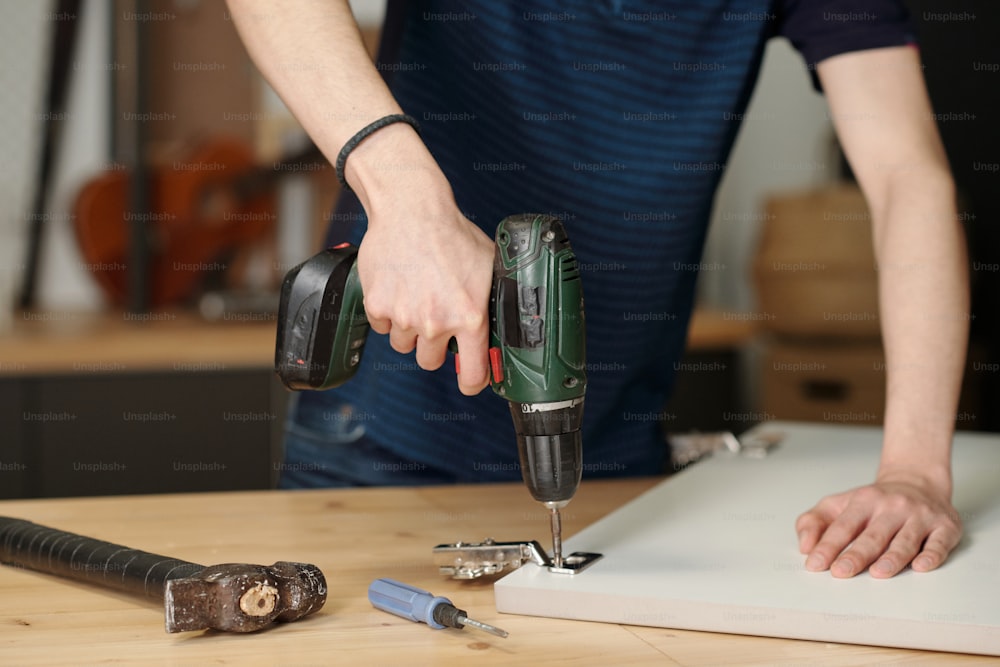 Hands of youthful boy holding electric drill while fixing hinge on door of kitchen cabinet while standing by workbench in garage