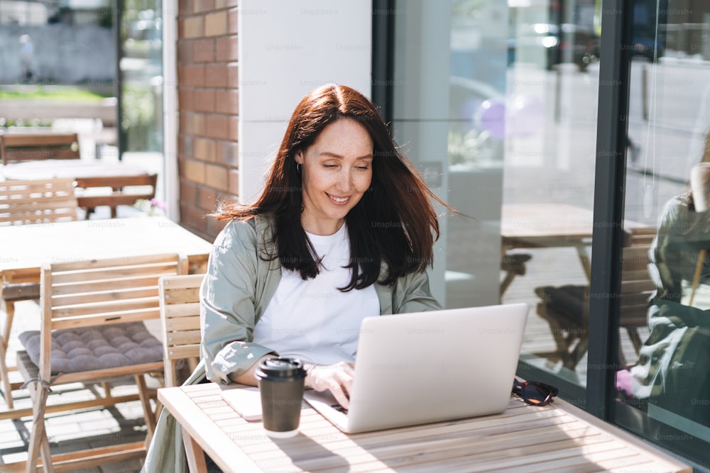 Femme d’affaires brune souriante adulte quarante ans en chemise élégante travaillant sur un ordinateur portable dans un café à la rue de la ville