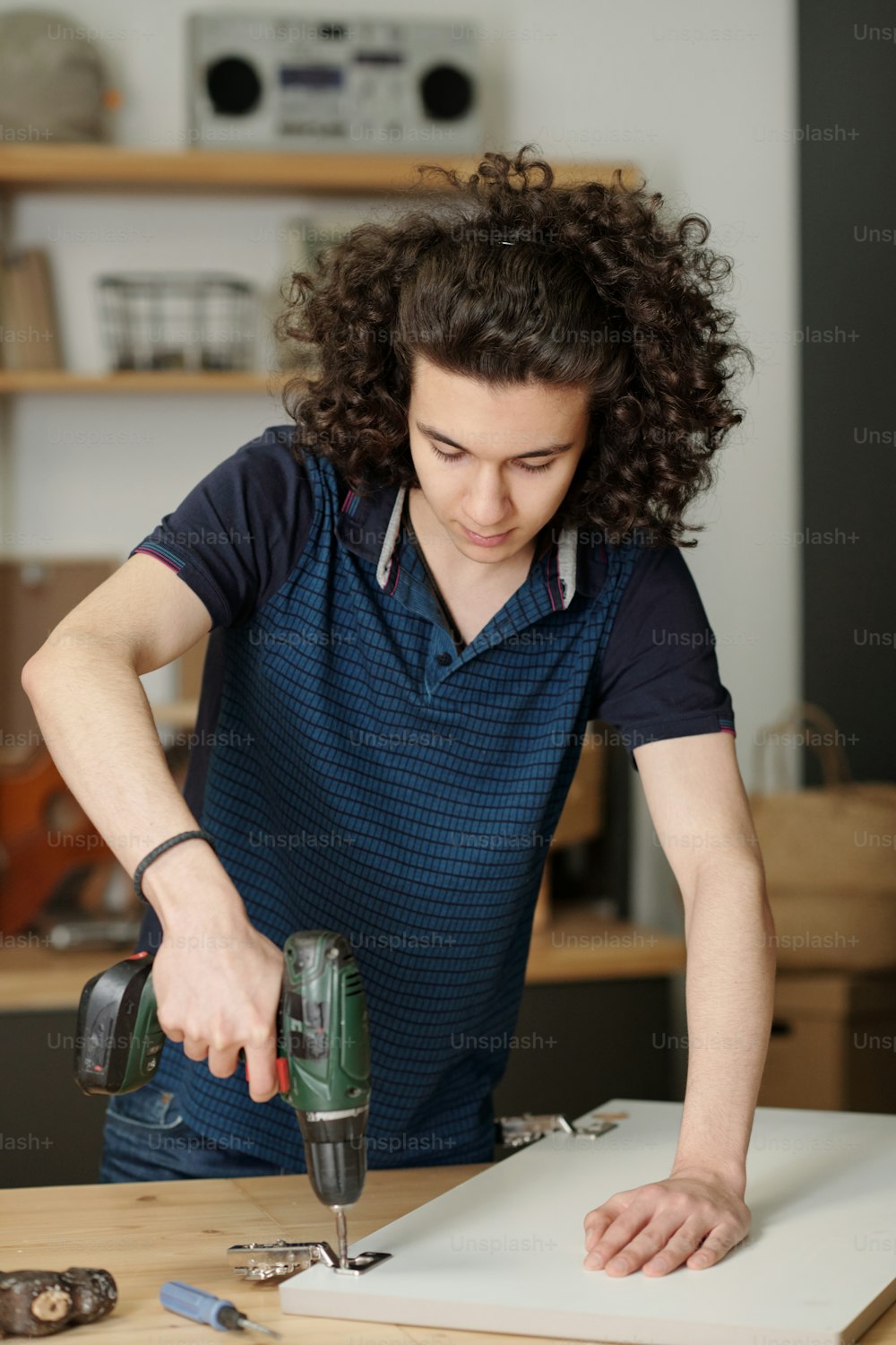 Jugendlicher Typ mit dunklem welligem Haar, das Sperrholztür des Küchenschranks bohrt, während er es von der Werkbank in der Garage seines Hauses repariert