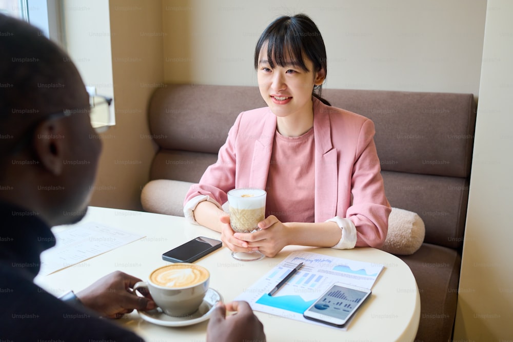Asian young woman drinking coffee and consulting with financial expert during meeting at cafe