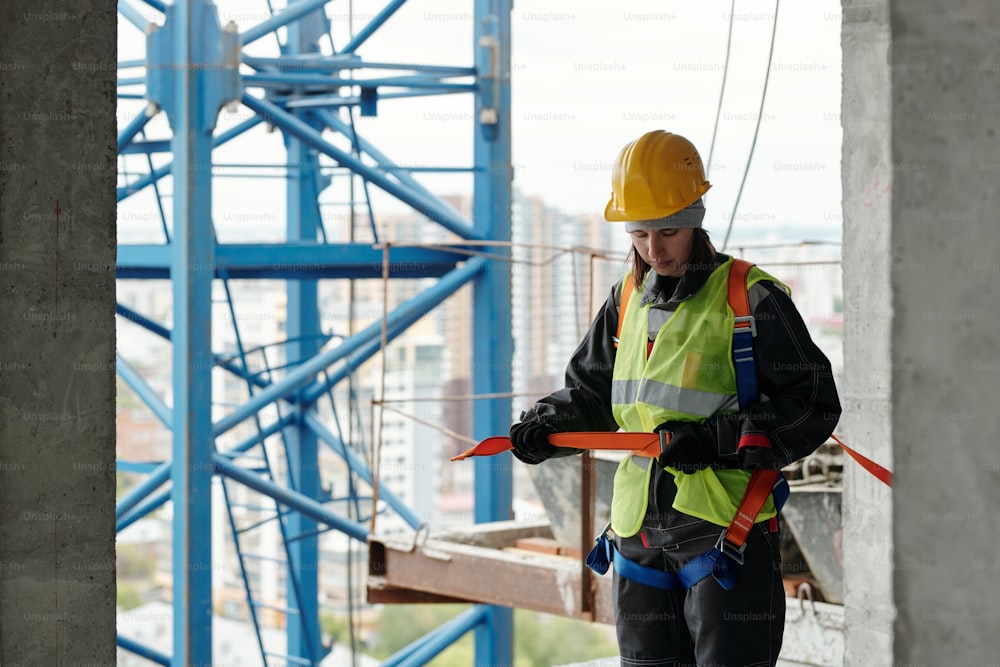 Mujer joven con casco protector y chaleco reflectante que sujeta el cinturón de seguridad en su cintura mientras va a trabajar en el sitio de construcción