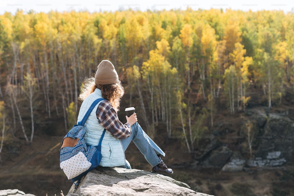 Young beautiful woman with curly hair in plaid shirt, jeans looks at the magic view of mountains and river, hiking on autumn nature