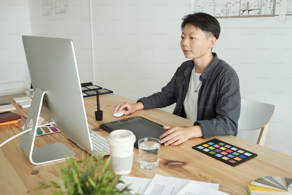 Young Chinese female designer sitting in front of computer monitor by desk in office