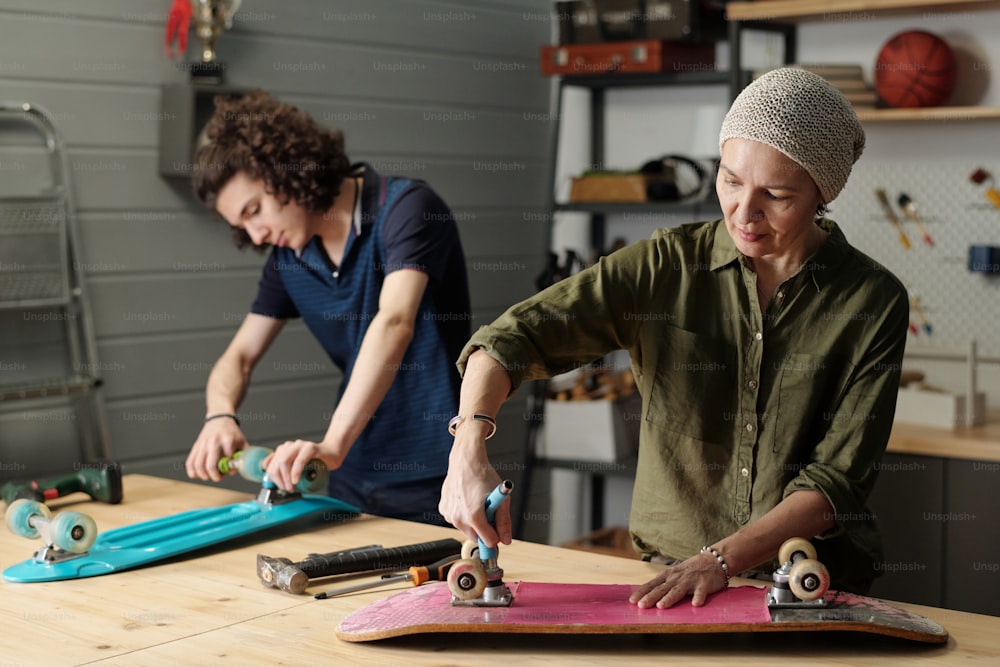 Zeitgenössische reife Frau und ihr jugendlicher Enkel reparieren Skateboards in der Garage, während sie am Holztisch stehen