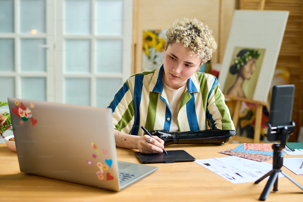 Joven diseñador gráfico y bloguero con prótesis de brazo creando un nuevo dibujo mientras mira la pantalla de la computadora portátil frente a la cámara del teléfono inteligente
