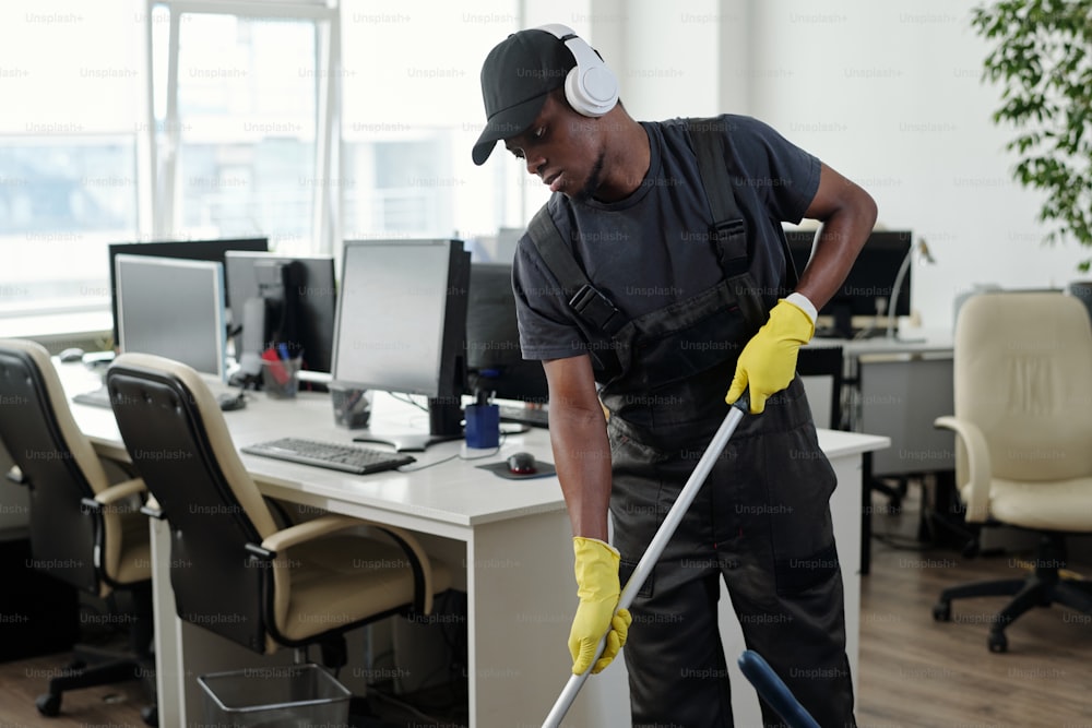Young male staff of cleaning service company in headphones washing floor in large openspace office and listening to music