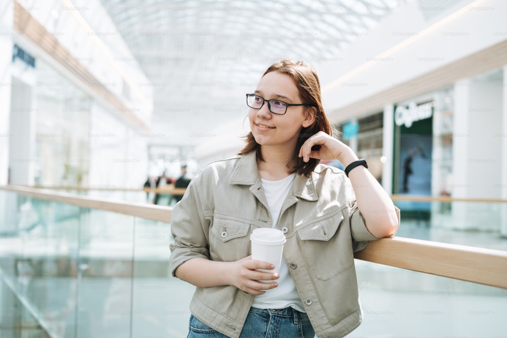 Junge brünette lächelnde Teenager-Studentin in Gläsern mit Pappkaffeebecher im öffentlichen Einkaufszentrum