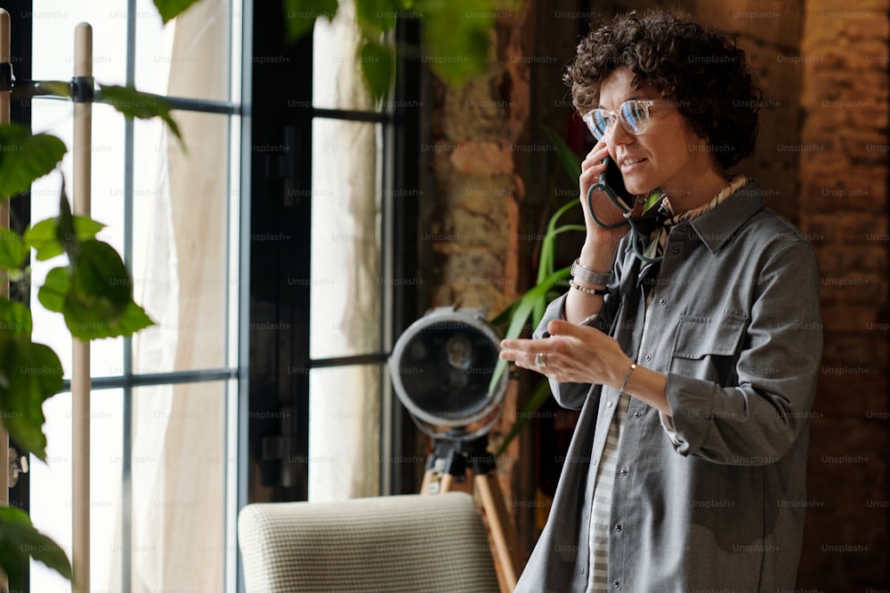 Junge Managerin oder Besitzerin eines Cafés in Freizeitkleidung, die mit Kunden telefoniert, während sie vor einem großen Fenster steht