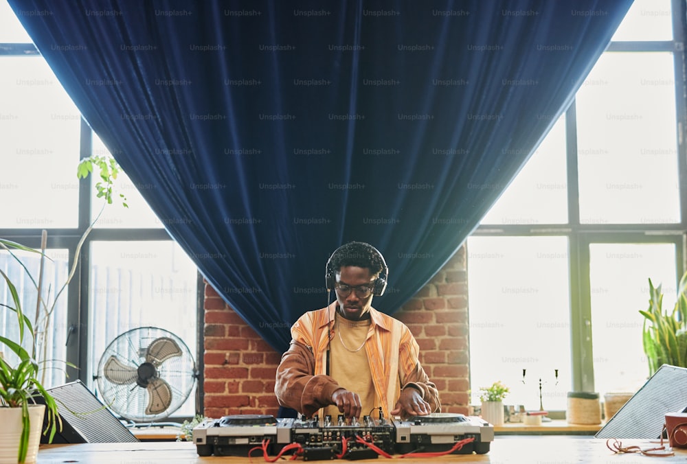 ヘッドフォンを着けた若い黒人男性が、DJコントローラーの前で新しい音楽を作りながら、サウンドミキサーを回して調整しています