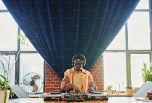 Joven hombre negro con auriculares creando nueva música frente al controlador de dj mientras gira y ajusta los mezcladores de sonido