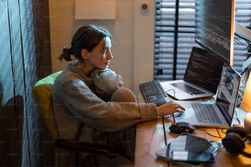 Young focused woman feeling overworked and tired, working on laptop and desktop computers, sitting at home office during the nighttime. Female programmer writing code