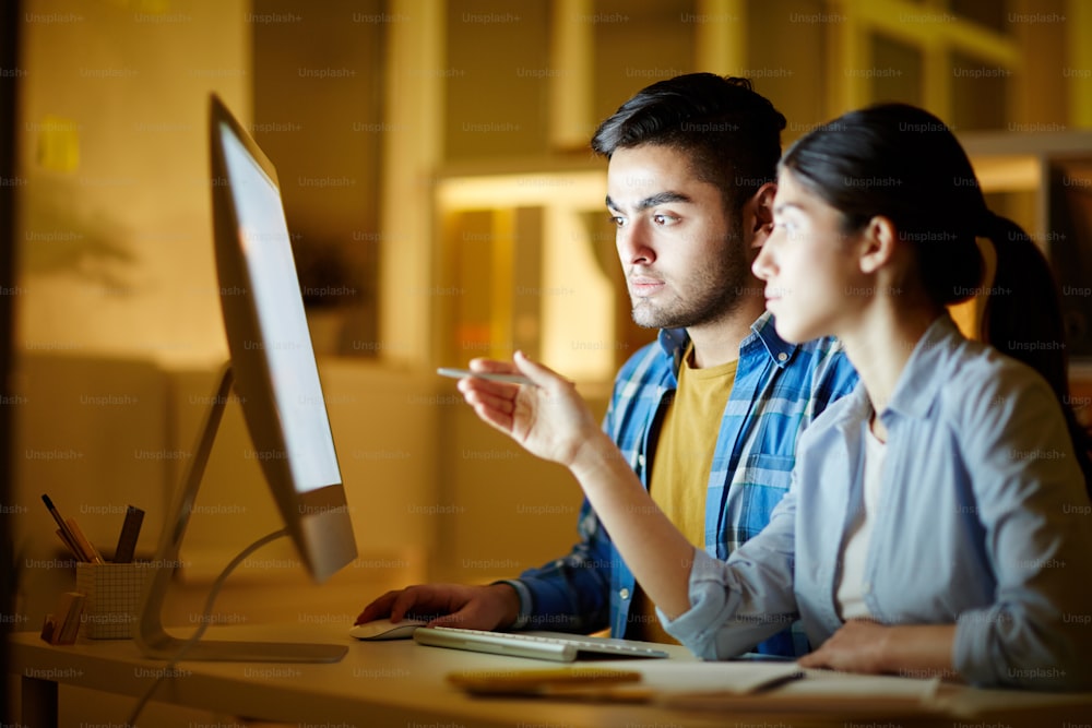 Young woman pointing at monitor display and showing her colleague new online project or course for beginners in sphere of web design