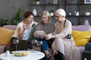 Mature women watching old photos together on the sofa, they drinking wine and remembering past moments during meeting at home