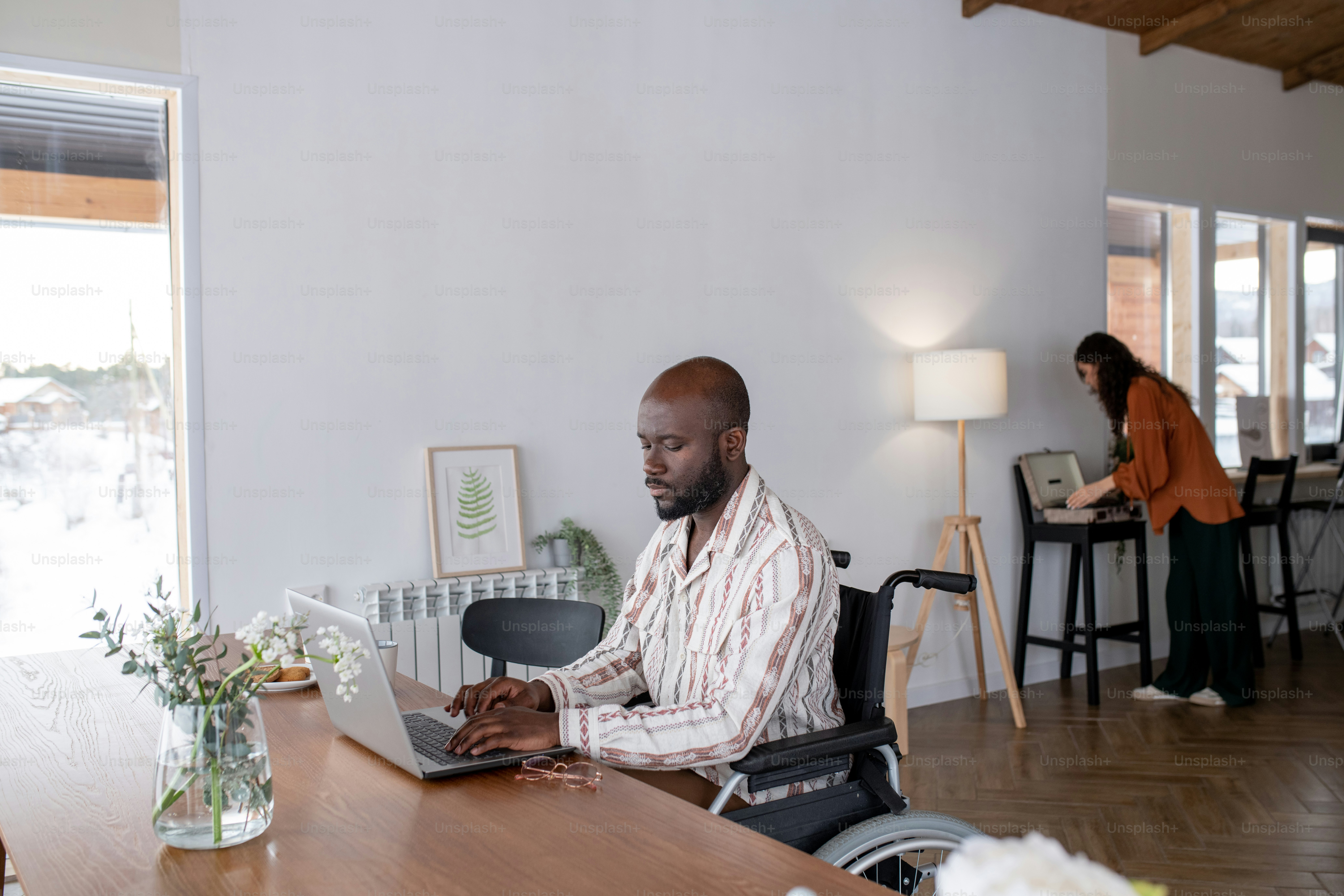 Hombre afroamericano serio en silla de ruedas sentado junto a la mesa frente a la computadora portátil y la red contra la mujer que enciende el tocadiscos