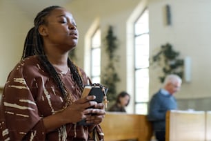 Jovem mulher negra em vestido casual mantendo os olhos fechados durante a oração silenciosa na igreja enquanto segurava a Bíblia Sagrada e as contas do rosário