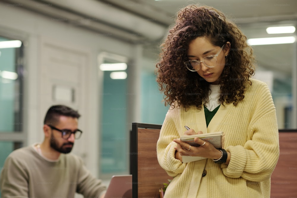 Young serious businesswoman in eyeglasses making notes of plan for the day while standing against colleague networking in front of laptop