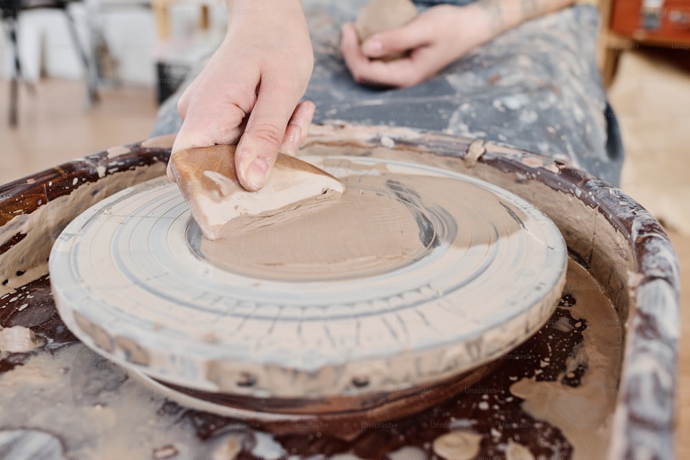 Mão do oleiro feminino criativo jovem aplicando argila na roda de cerâmica rotativa com esponja molhada antes de criar novo item de barro