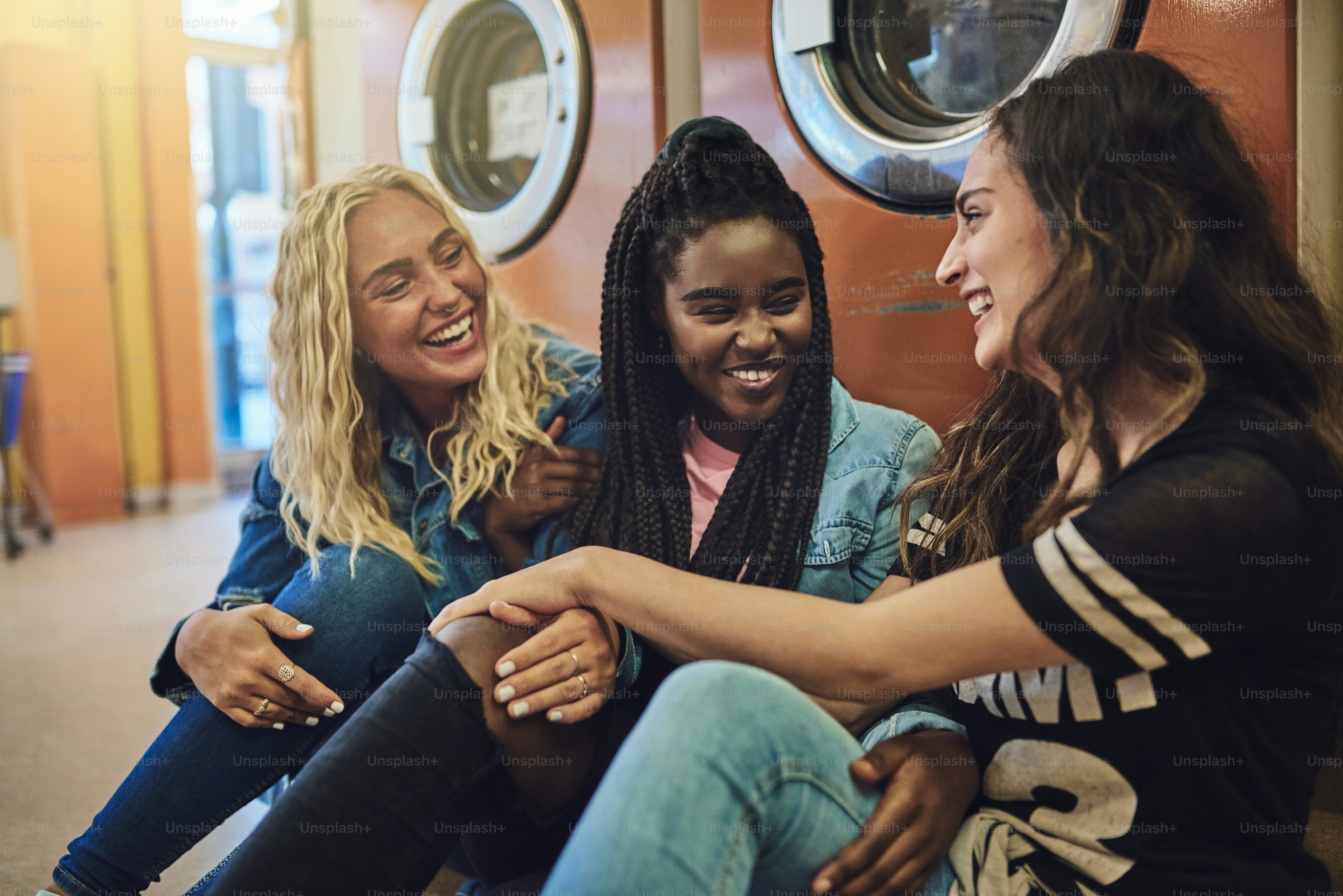 Groupe diversifié de jeunes amies assises sur le sol d’une laverie automatique en train de parler et de rire ensemble tout en faisant la lessive