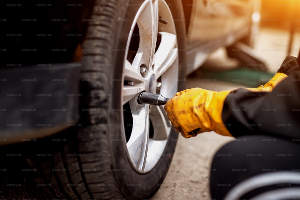 An experienced hard working mechanic in orange gloves is using an electric wrench to tighten up the screws in a car wheel.