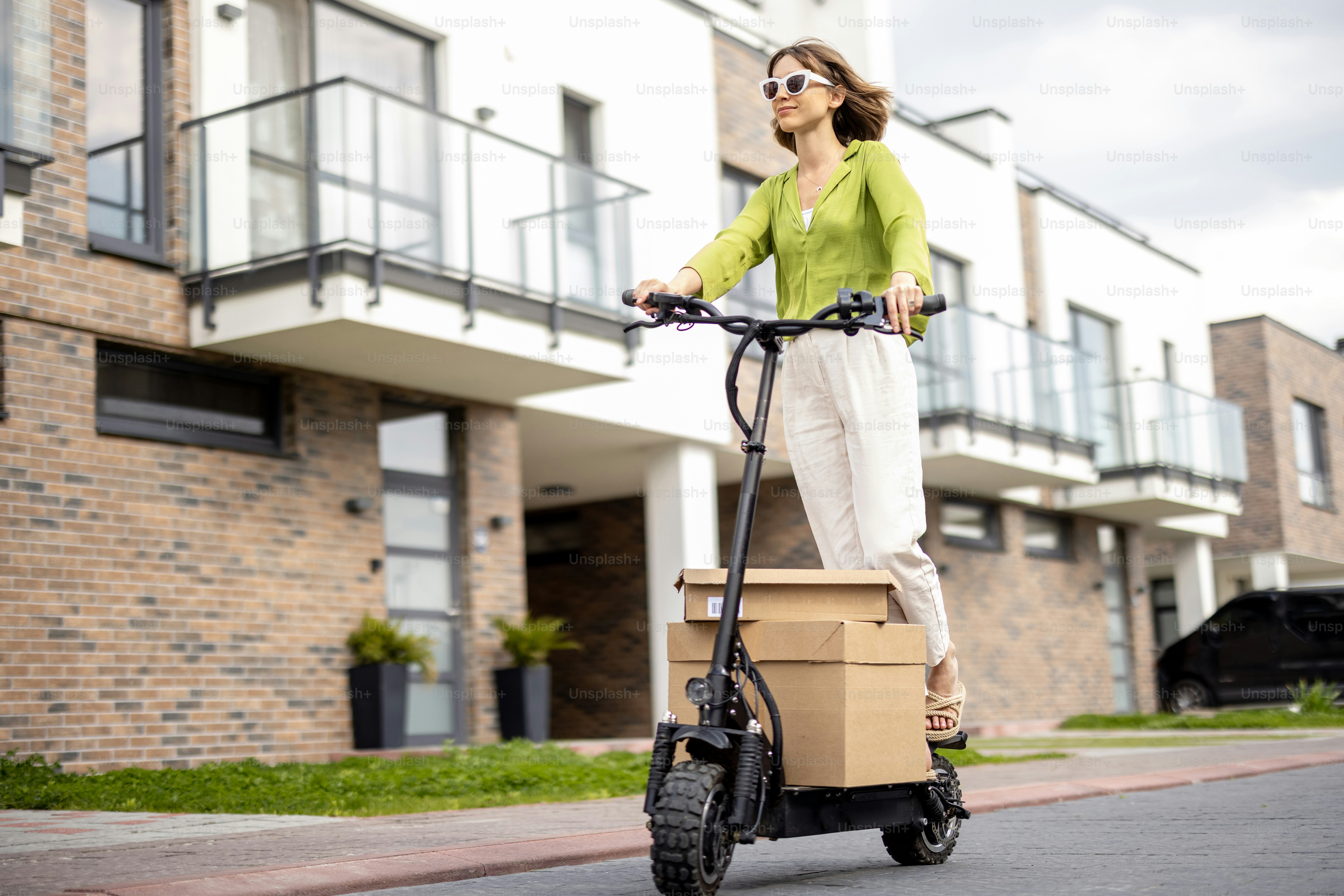 Young happy woman driving electric scooter, carrying cardboard boxes on street in residential area. Concept of sustainability, delivery and eco-friendly modern lifestyle