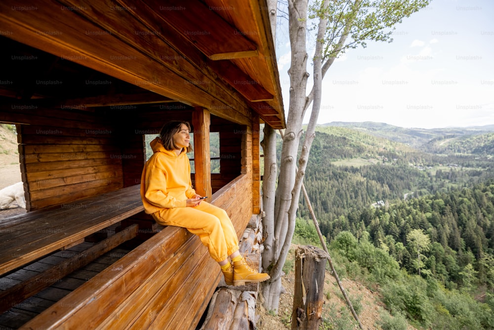 Young woman in orange sport suit enjoys great mountain landscape while sitting on window of wooden tiny house. Concept of escape to nature and traveling in the mountains
