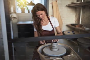 도자기 스튜디오에 앉아 도자기 바퀴를 돌리는 점토 조각을 창의적으로 만들기 위해 조각 도구를 사용하는 여성 장인