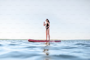 Mulher jovem bonita em paddleboarding maiô preto no lago com ondas durante a luz da manhã