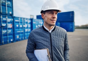 Ingeniero sonriente con un casco y sosteniendo un portapapeles mientras está solo en un gran muelle de envío comercial