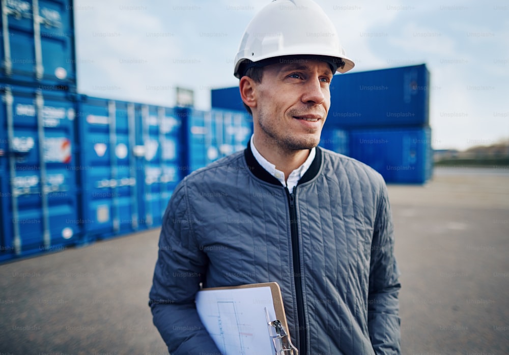 Smiling engineer wearing a hardhat and holding a clipboard while standing alone on a large commercial shipping dock
