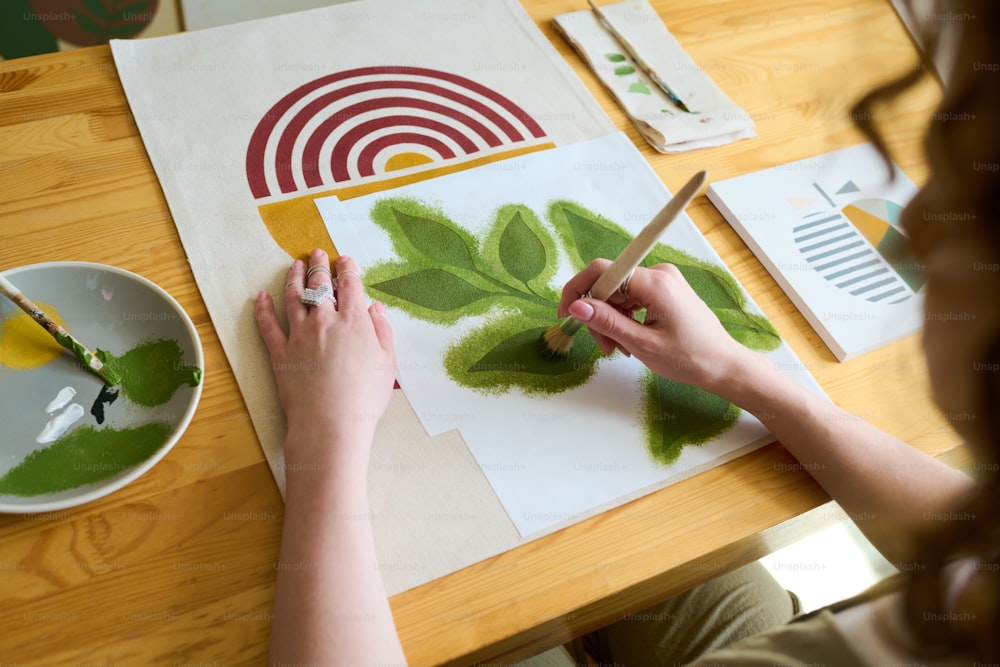 Hände einer jungen Studentin der Kunsthochschule, die grüne Blätter mit Pinsel und Gouache auf Papier malen, während sie am Tisch sitzt