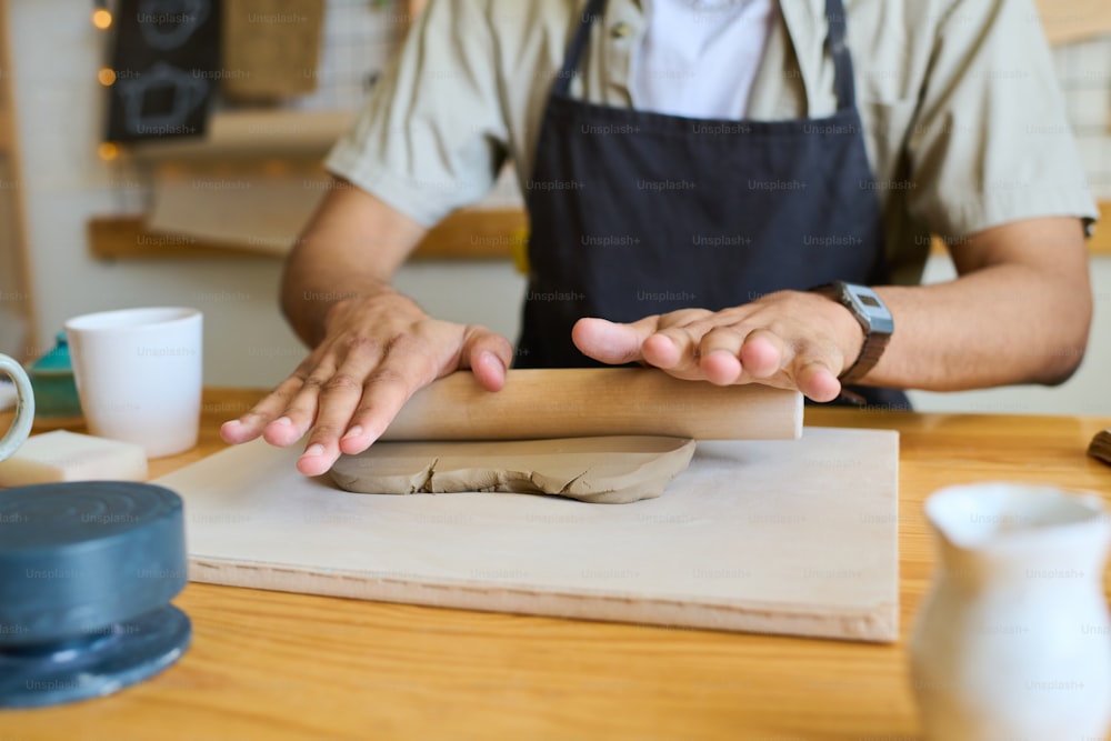 Hands of young black man in apron flattening piece of clay with rolling pin on board while sitting by table with supplies for handwork