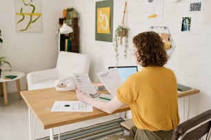 Back of young female employee in casualwear comparing two financial documents while looking through data by workplace at home