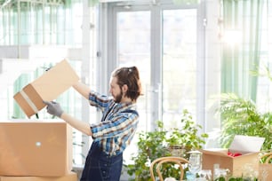 Serious busy bearded young moving company worker picking up box and reading label while getting information about delivered goods in modern space with plants