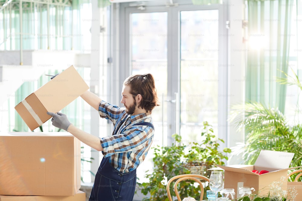 Serious busy bearded young moving company worker picking up box and reading label while getting information about delivered goods in modern space with plants
