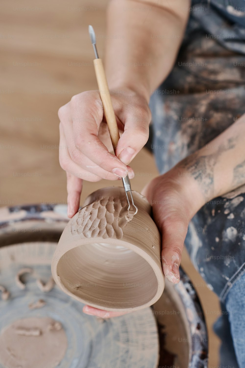 Hands of young creative female ceramist decorating handmade clay cup with carved patterns in workshop while using sharp handtool