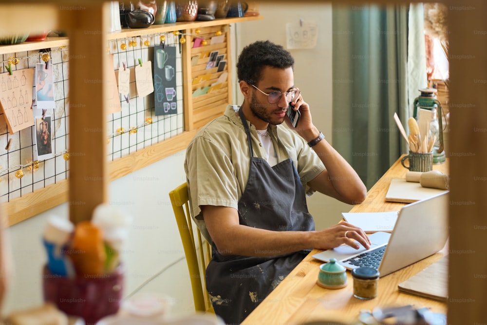 Funcionário do sexo masculino jovem no avental chamando clientes e olhando através de pedidos on-line enquanto sentado no local de trabalho na frente do laptop