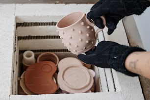 Hands of young female in black gloves taking handmade clay cup out of square container with other earthenware items made for sale