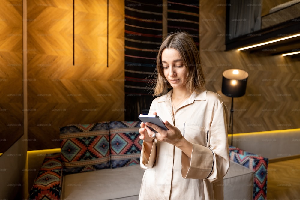 Woman using smart phone while standing in living room of a modern style apartment. Concept of online communication and home comfort. Caucasian woman wearing beige sleepwear