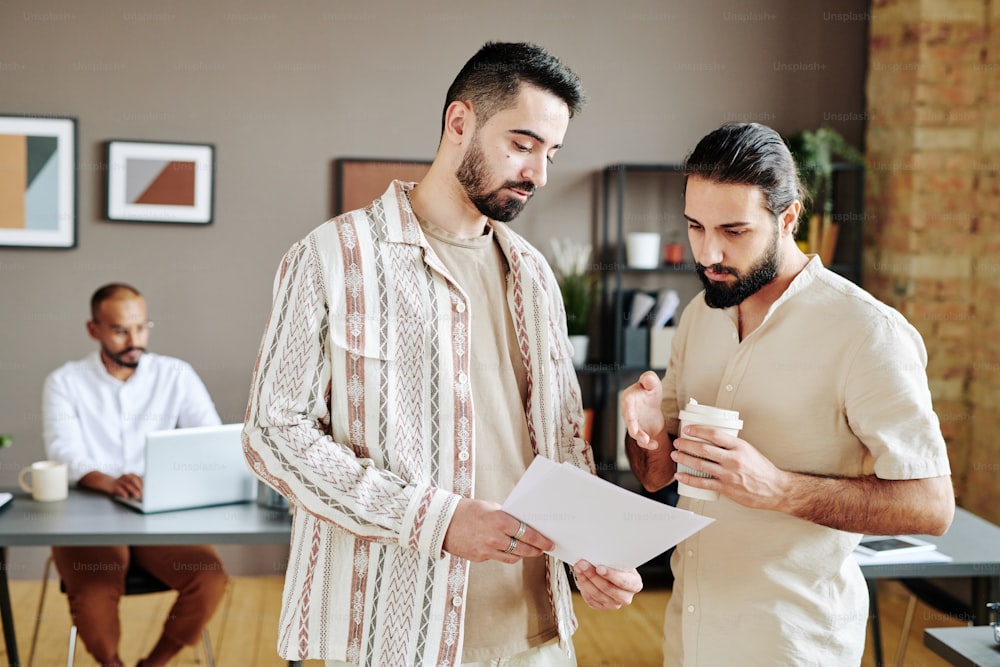 Young confident businessman showing his colleague or ceo financial paper and discussing information with him at meeting in office