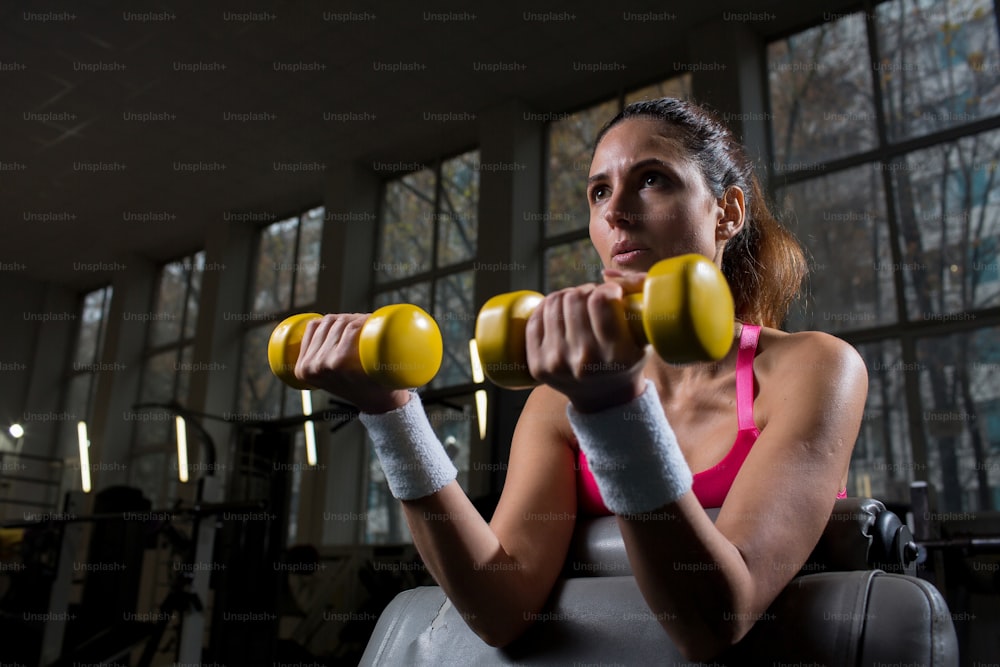 Starke junge Frau mit gelben Hanteln, die Übungen im Fitnessstudio macht