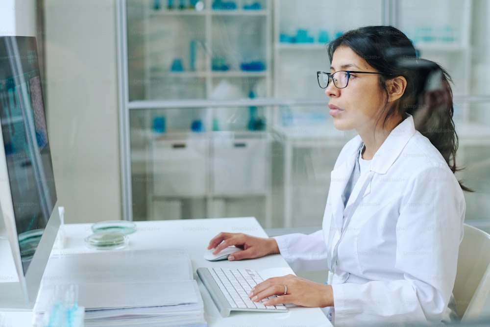 Young pretty female scientist in whitecoat and eyeglasses using computer for studying new virus while sitting by workplace in laboratory