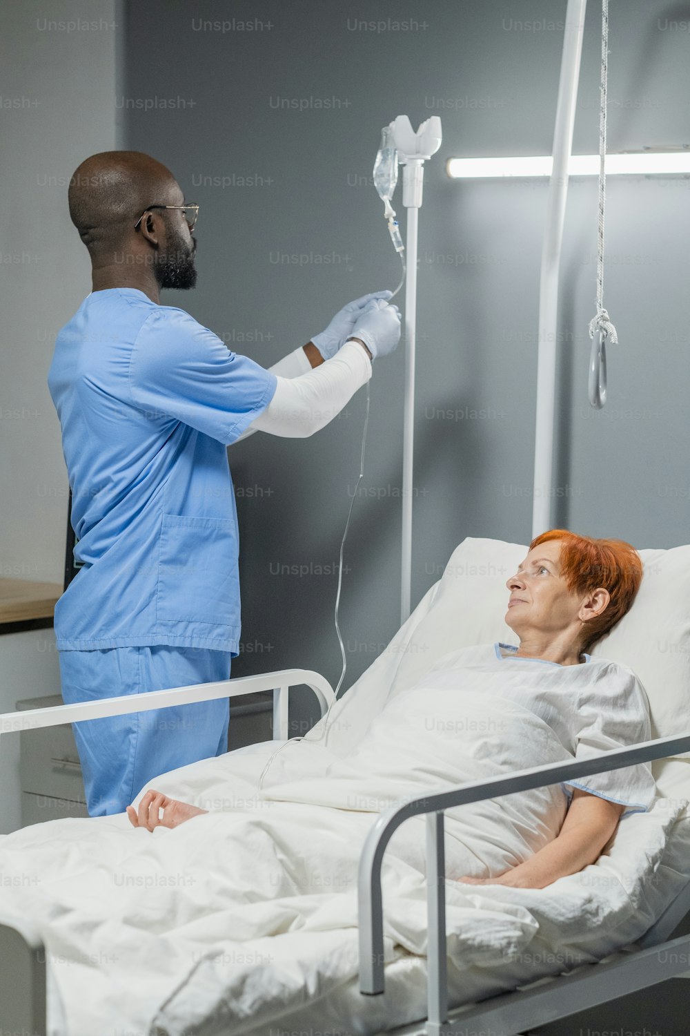 African anesthesiologist in uniform making dropper for sick senior woman while she lying in bed at hospital