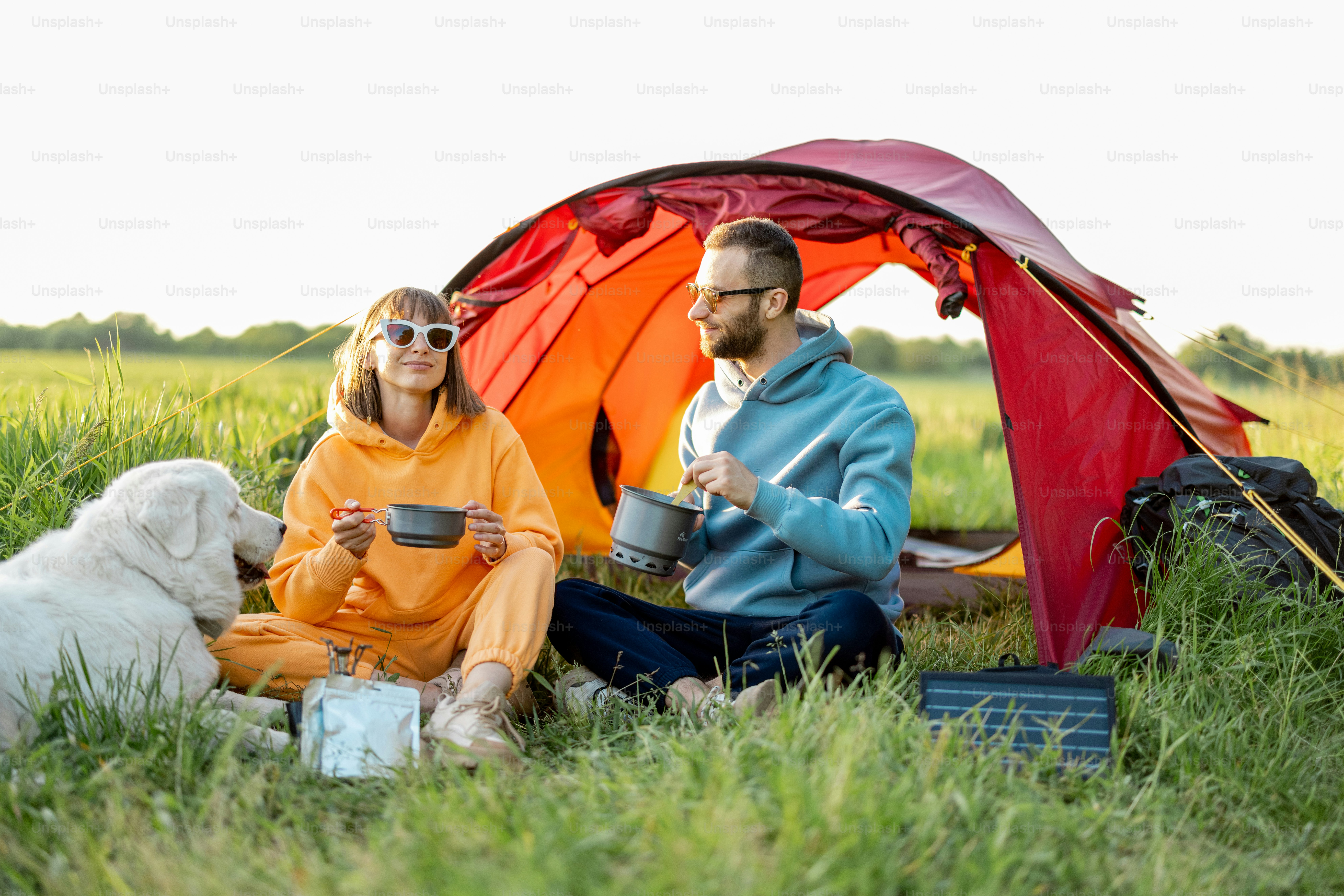 Young couple cooking food, spending summer time with a dog at campsite on the green field. Man and woman travel with tent on nature